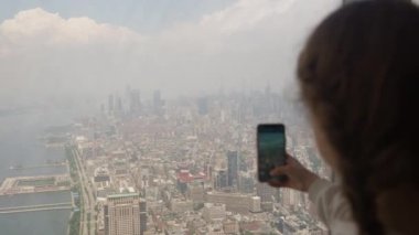 Medium Slow Motion Handheld Shot Of Girl Filming With Smartphone Through Window In Skyscraper To New York Cityscape Below