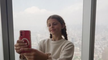Medium Slow Motion Handheld Shot Of Girl Posing For Selfie With Smartphone And Arranging Plaits By Window In Skyscraper With New York City Below