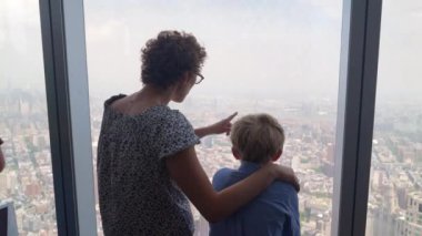 Medium Slow Motion Handheld Shot Of Mother With Arm Around Young Boy Looking Through Skyscraper Window To New York City Below