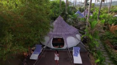 Aerial View of a Luxury Resort Tent House Between Coconut Trees in Tropical Forest. Pretty Woman Gets Off the Tent and Walks Along the Pathway. Tilt Down Zoom Out Shot
