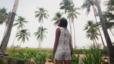 Pretty Asian Woman Stands on a Terrace of a Tropical Resort and Enjoys the View of Coconut Trees in Front of Her. Tilt Up Arc Shot