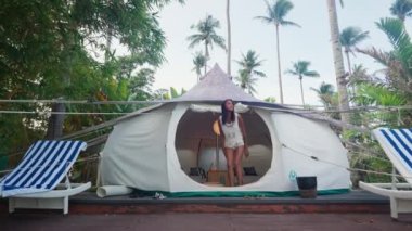 Pretty Asian Woman Gets Out of a Tent House to Terrace. Luxury Tropical Resort Between Coconut Trees. Dolly Arc Shot. Camera Follows the Woman