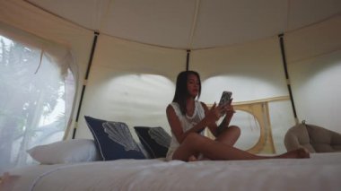 Pretty Asian Woman Sitting on the Bed Inside Luxury Tent House and Using Her Mobile Phone. Static Shot