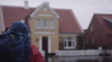 Close-up Back View of a Man with Backpack in Winter Clothes Walking Along the Road Between Residential Houses at the Rain. Bokeh Slow Motion Shot