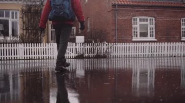 Man with Backpack in Winter Clothes Walking on a Wet Asphalt Road with Reflections on Surface at the Rain. Slow Motion Tilt Up Shot