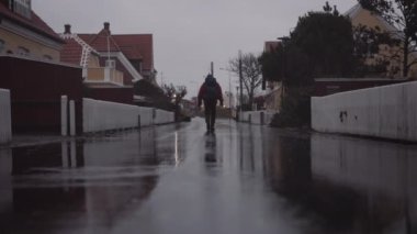 Man with Backpack Walking on a Wet Asphalt Road with Reflections on Surface Between Residential Houses at the Rain. Slow Motion Static Shot