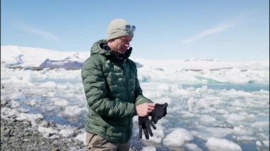 Man Tourist Puts On His Gloves in Front of Jokulsarlon Glacier Lagoon, Iceland. Slow Motion Panning Shot