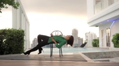 Wide Dolly Slow Motion Shot Of Dancer In Green And High Heels Bending Over Backwards On Chair By Infinity Pool In City