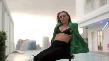 Zoom In Slow Motion Medium Shot Of Sensuous Dancer Crossing Legs In Green Jacket And Bra On Chair And Looking At Camera