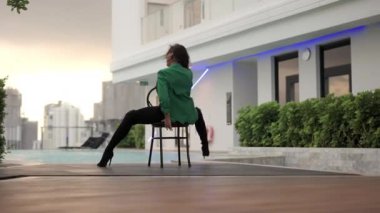 Slow Motion Dolly Wide Shot Of Young Dancer In Green Jackets And High Heels Sitting In Chair Then Turning To Side
