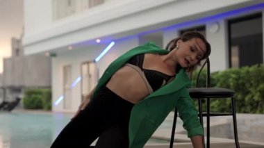Medium Dolly Slow Motion Shot Of Sensuous Dancer Posing By Chair At Poolside In Green Jacket And Bra