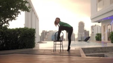 Wide Slow Motion Lockdown Shot Of Dancer In High Heels And Green Jacket Sitting On Chair At Poolside With Back To Camera