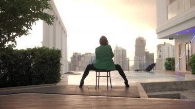 Wide Slow Motion Lockdown Shot Of Dancer In High Heels And Green Jacket Sitting And Dancing On Chair At Poolside With Back To Camera