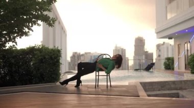 Wide Slow Motion Lockdown Shot Of Dancer In High Heels And Green Jacket Sitting And Bending Over Backwards On Chair At Poolside