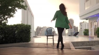 Wide Lockdown Shot Of Empty Chair At Poolside As Woman In Green Jacket In High Heels Walks Towards Pool