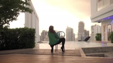 Wide Lockdown Slow Motion Shot Of Sensuous Woman In Bra And High Heels Crouching Then Standing And Turning To Pose On Chair At Poolside