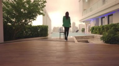 Wide Slow Motion Tracking Shot Of Woman In Green Jacket And High Heels Walking To Chair At Poolside Then Turning To Camera