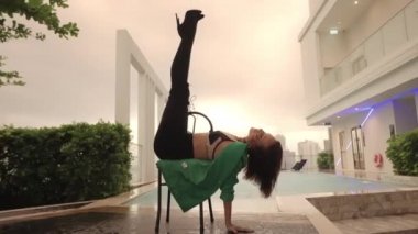 Wide Slow Motion Dolly Shot Of Dancer In High Heels And Green Jacket Leaning Back And Kicking Legs In Air On Chair At Poolside
