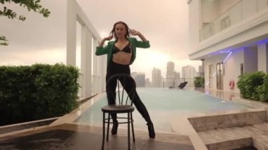Medium Slow Motion Dolly Shot Of Dancer Performing In Bra And Green Jacket At Poolside With Chair