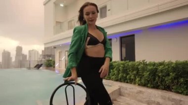 Medium To Wide Slow Motion Dolly Shot Of Dancer Performing In Bra And Green Jacket At Poolside With Chair