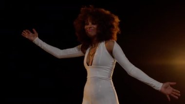 Smiling African American Woman with Afro Style Hairs in White Jumpsuit Dancing on the Stage  Against Dark Background and Stage Light. Cinematic Close-up Slow Motion Zoom In and Zoom Out Shot