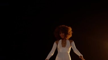 Smiling African American Woman with Afro Style Hairs in White Jumpsuit Dancing on the Stage Against Dark Background and Stage Lights. Cinematic Slow Motion Shot