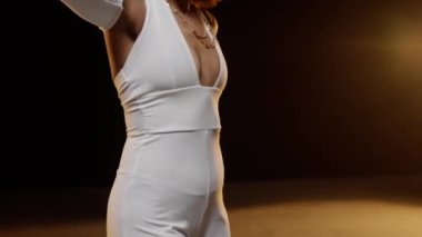 Portrait of Smiling African American Woman with Afro Style Hairs in White Jumpsuit Dancing on the Stage Against Dark Background and Stage Lights. Cinematic Slow Motion Shot