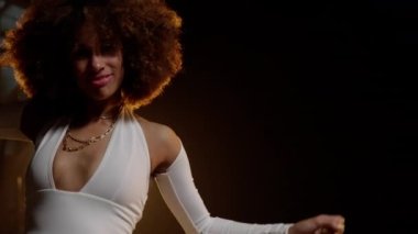 Portrait of Smiling African American Woman with Afro Style Hairs in White Jumpsuit Dancing on the Stage Under Shiny Disco Ball. Cinematic Slow Motion Shot. Camera Follows the Woman