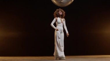 Smiling African American Woman with Afro Style Hairs in White Jumpsuit Wearing Roller Scates Dancing on the Stage Under Shiny Disco Ball. Cinematic Slow Motion Zoom In Shot