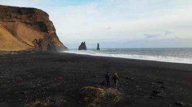 Wide Drone Rising Over Two Drone Pilots Standing On Black Sand Beach In Spring, Iceland