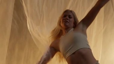 Close-up of a Professional Dancer Performing in Studio in Soft Warm Lighting. Graceful Young Woman with Long Hairs Dancing with White Stage Curtain. Slow Motion Shot