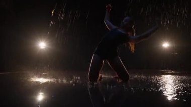 Sensual Young Woman Dancer Performing in the Rain in Dark Studio Against Black Backdrop and with Spotlights On. Water Drops Falling on the Floor Imitating Rain Shower. Dolly In and Out Shot
