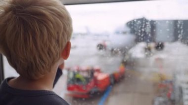 Orta şiddette ağır çekimde, yağmurda pistte havaalanında pencereden dışarı bakan genç bir çocuk görülüyor.