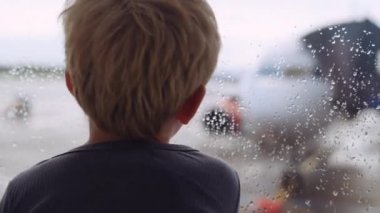 Orta şiddette ağır çekimde, yağmurda pistte havaalanında pencereden dışarı bakan genç bir çocuk görülüyor.