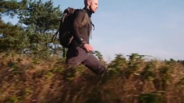 Bald Head Turist Sırt Çantası Yürüyüşleri Ormanda Uzun Çimenler Çayırında. Hillock 'tan atlar ve ileriye doğru yürür. Kamera Adamı Takip Ediyor