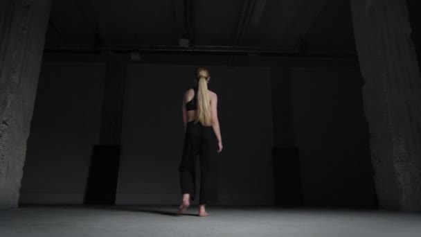 精力充沛的年轻女舞蹈家 金发碧眼 头戴黑色上衣 裤子与混凝土墙一起独自在工作室里跳舞 静态射击 — 图库视频影像