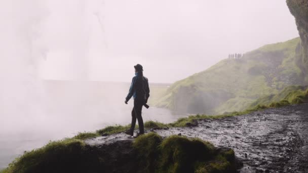 アイスランドのSeljalandsfoss滝の後ろに立つ男性写真家 彼は手を挙げている 水は力で落ちる 雄大な景色 スローモーションショット — ストック動画