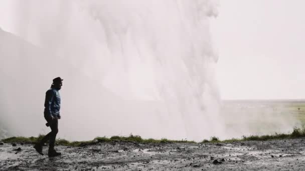 アイスランドのSeljalandsfoss滝の後ろを歩く男の写真家 水は力で落ちる 雄大な景色 スローモーションショット — ストック動画