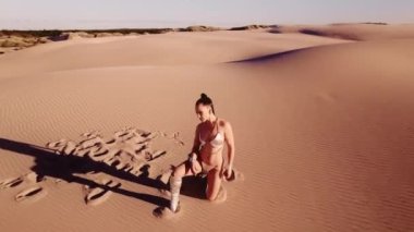 Geniş İHA Çölde Sıcak Oturan, Kum Kumu Giyen, Çizme Giyen ve Bikini Giyen Genç Kadın Üzerine Kavis Yapıyor