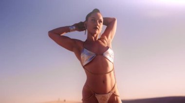 Gümüş Bikini giymiş, elleri çölde başının arkasında olan güzel bir kadının orta boy resmi.