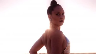 Gümüş bikinili, uzun örgülü saçlı, arkasında güneş ışığı olan genç bir kadının orta boy yavaş çekim görüntüsü.