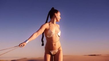 Gümüş bikinili ve çizmeli genç bir kadının el kamerasıyla çölde el çantasıyla dolaşırken çekilmiş.