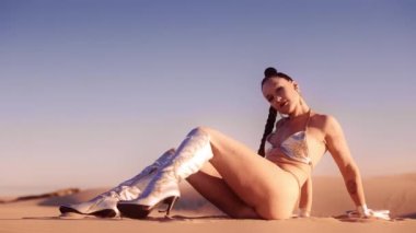 Geniş Dolly Ağır Çekim Gümüş Çizmeli Genç Kadın ve Bikini Oturuyor ve Çölde Poz Veriyor