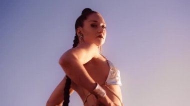 Düşük Açılı Yavaş Çekim: Gümüş Bikini giymiş, elleri kalçasında, mavi gökyüzüne bakan genç bir kadın.