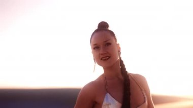 Gümüş bikinili, uzun örgülü saçlı, arkasında güneş ışığı olan genç bir kadının orta boy yavaş çekim görüntüsü.