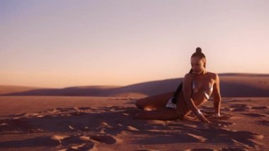 Gümüş Bikini giymiş, çölde yatıp gümüş çizmelerle kum tekmeleyen genç bir kadının ağır çekimleri.