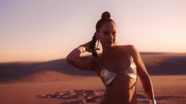 Gümüş Bikini giymiş, uzun örgülü saçlarını çölde sallayan genç bir kadının orta boy ağır çekim resmi.