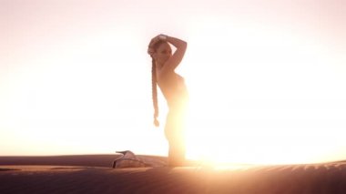 Gümüş bikinili ve çizmeli genç bir kadının güneşin altında çöl ortasında diz çöküp poz verişi.