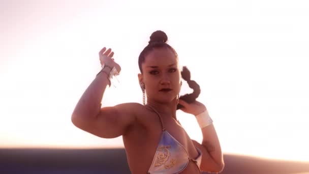 ロングヘアでシルバービキニトップの若い女の中出しスローモーション カメラを見て 彼女の後ろに日光で踊る — ストック動画