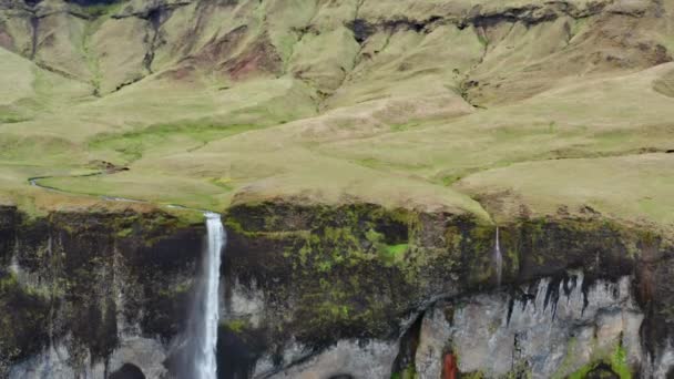 アイスランドの緑の風景から崖の上を流れる滝を下るワイドドローンショット — ストック動画